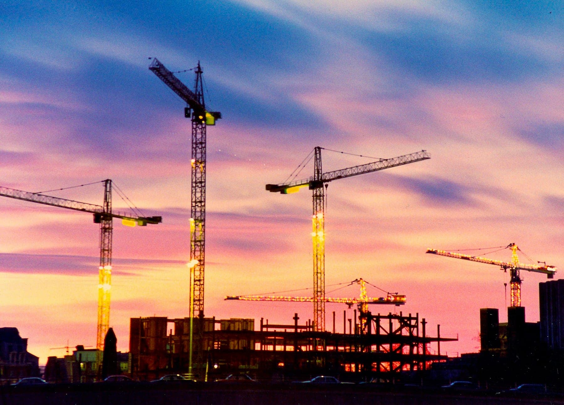 Tower cranes standing over a construction site, silhouetted against a sunset sky.
