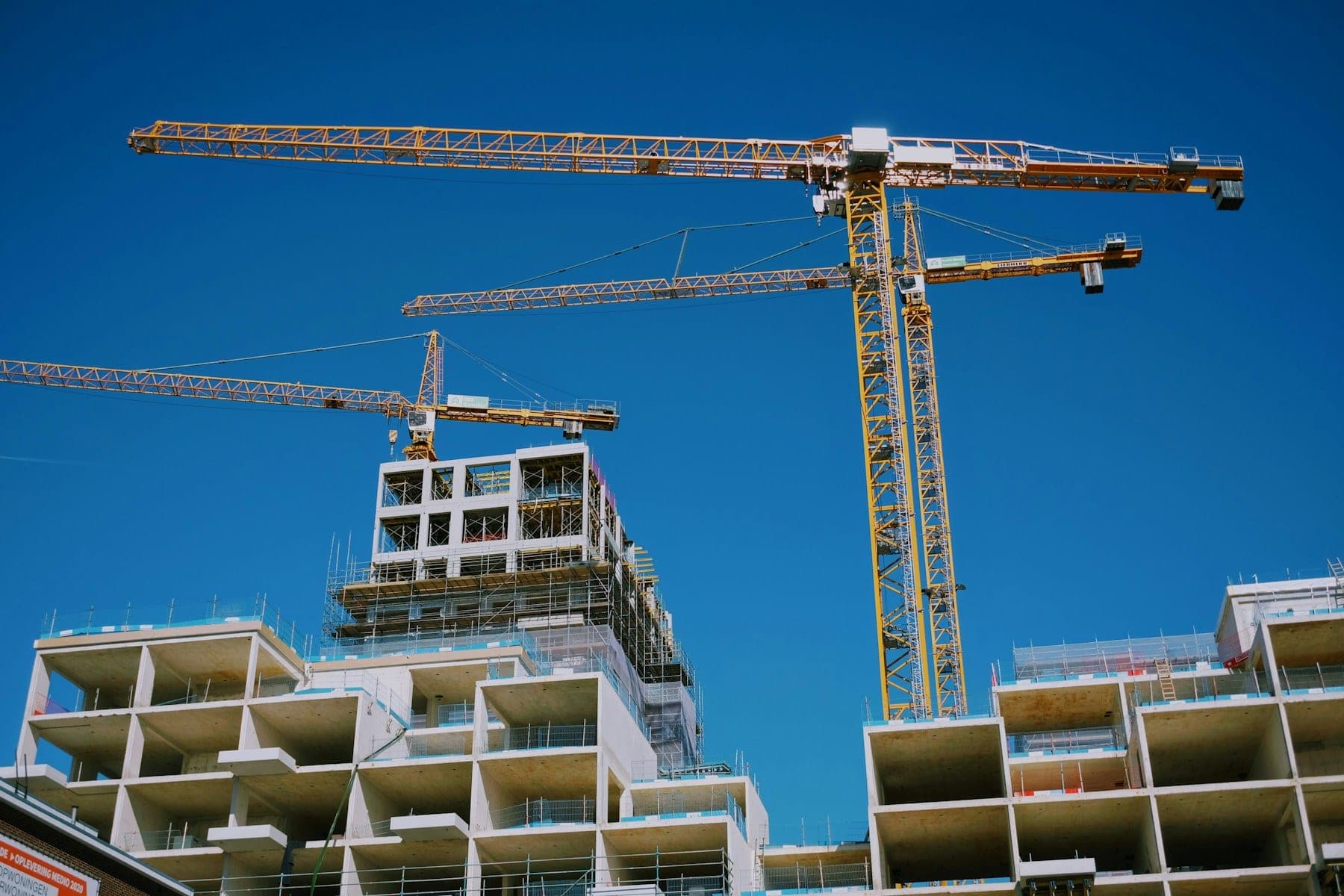 A multi-storey concrete building under construction, with tower cranes above it against a clear sky.