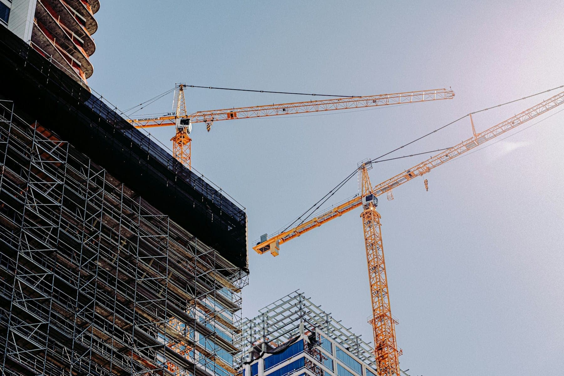 Tower cranes beside the steel frame and scaffolding of a building under construction, in low sunlight.
