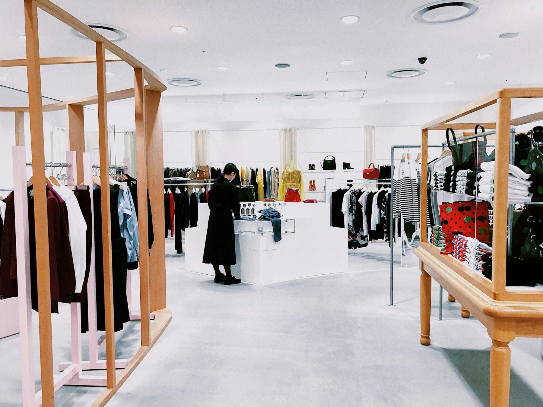 A bright, modern retail store floor: clothing rails, a central display table, and a shopper browsing the merchandise.