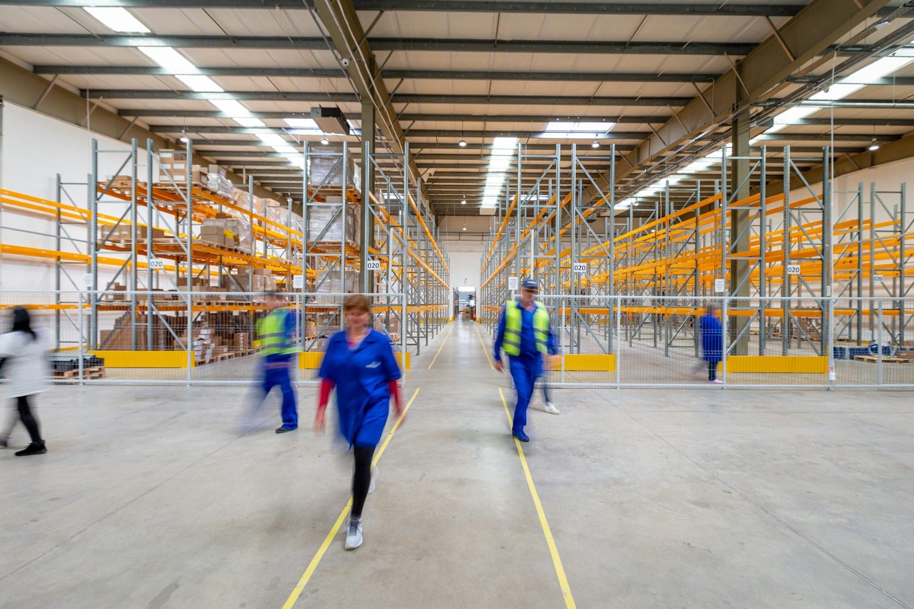 A fulfilment warehouse aisle lined with tall pallet racking, staff crossing the floor in motion blur.