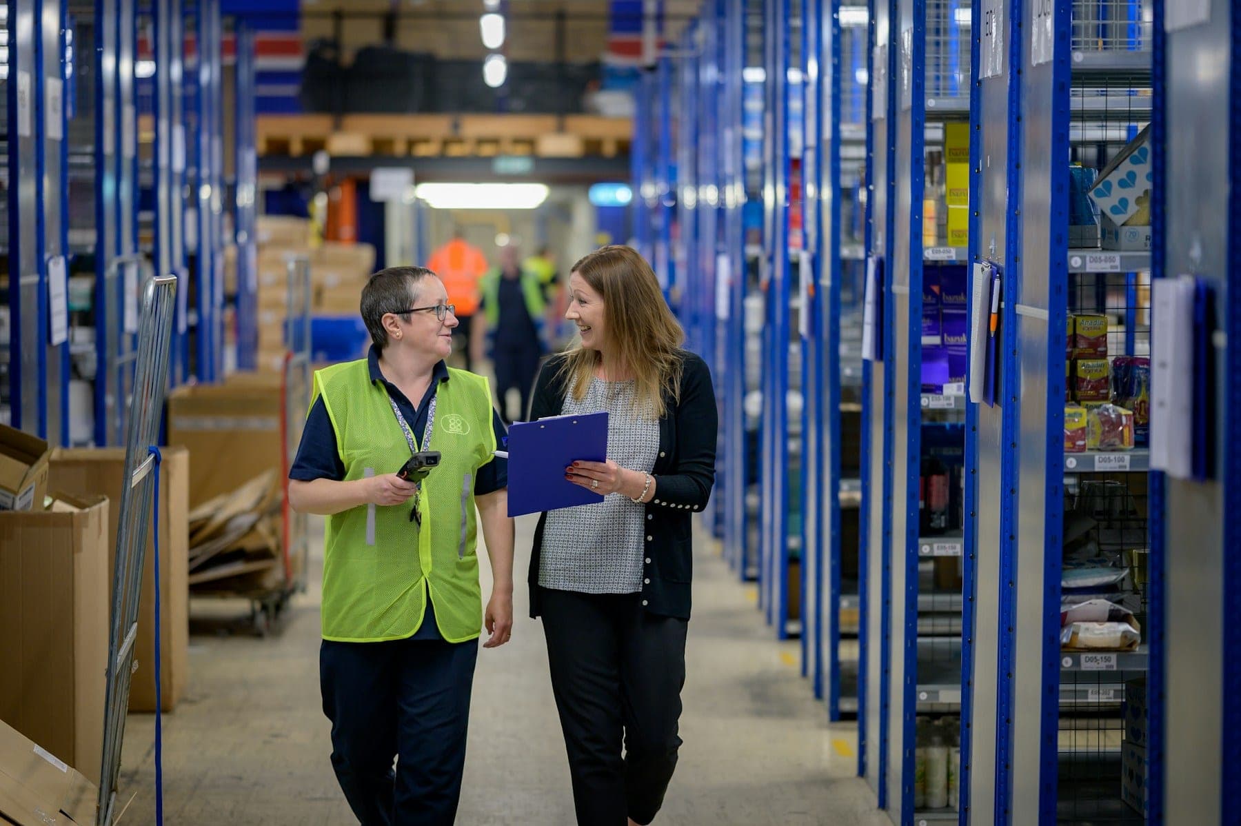 Two warehouse staff walking a stock aisle between blue racking, one holding a barcode scanner and the other a clipboard.