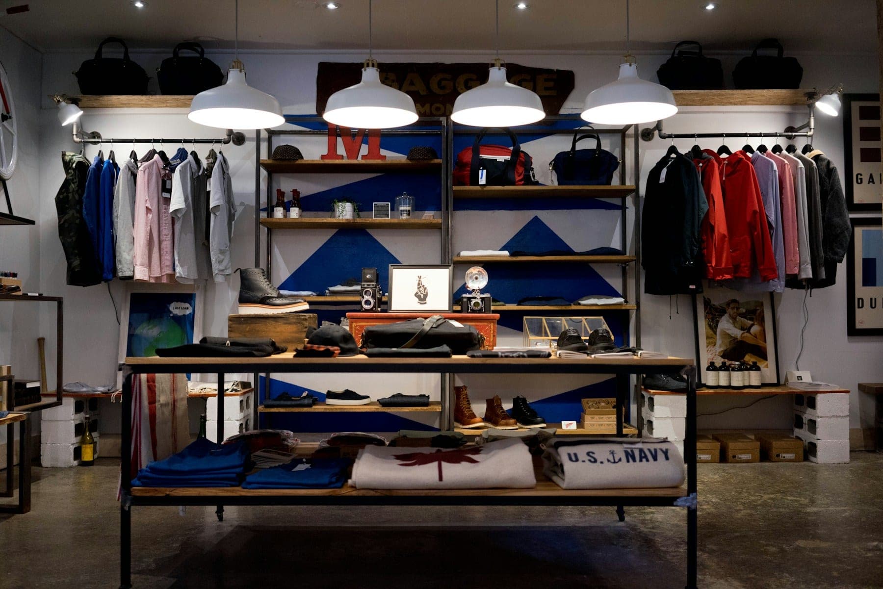 A boutique display table and shelves styled with folded garments, bags, and shoes under pendant lighting.