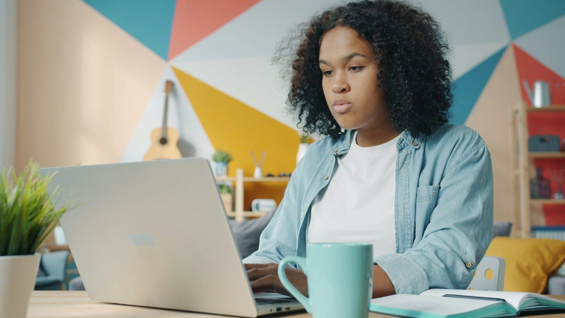 A young woman working on a laptop at a desk, an open notebook beside her, in a brightly decorated room.