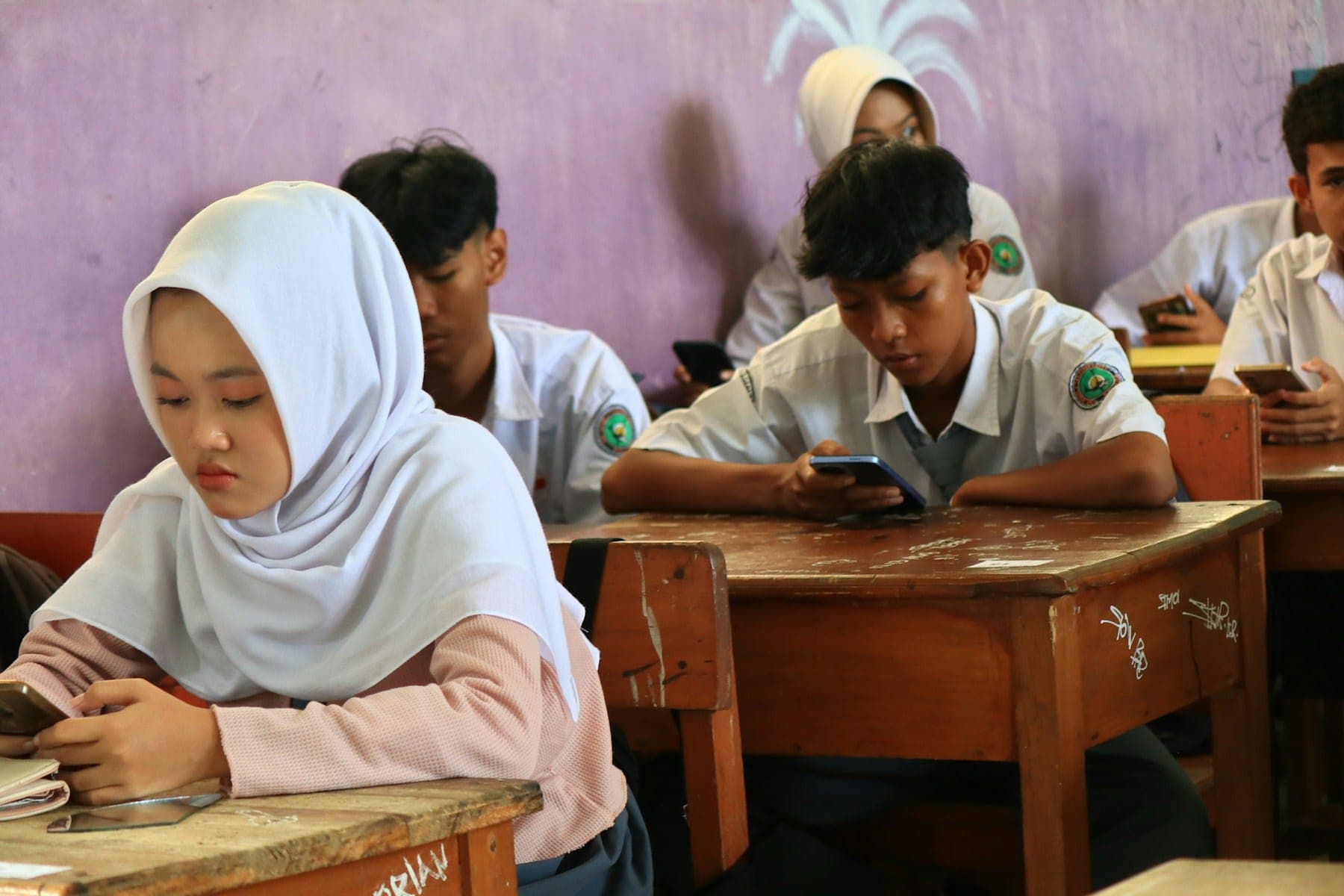 Students at wooden desks in a classroom, several of them using mobile phones.