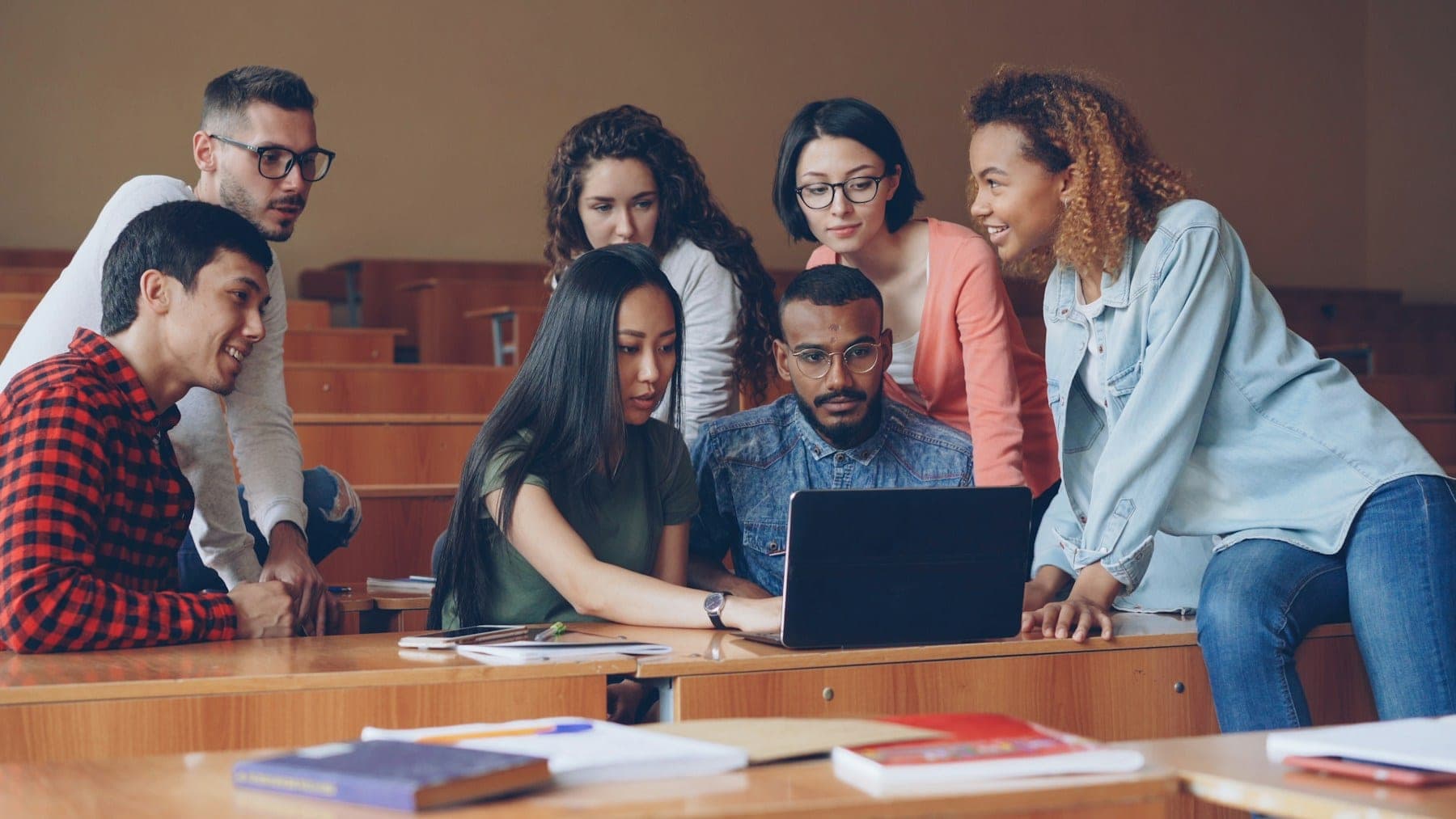 A group of students gathered around a laptop at a desk in a lecture hall.