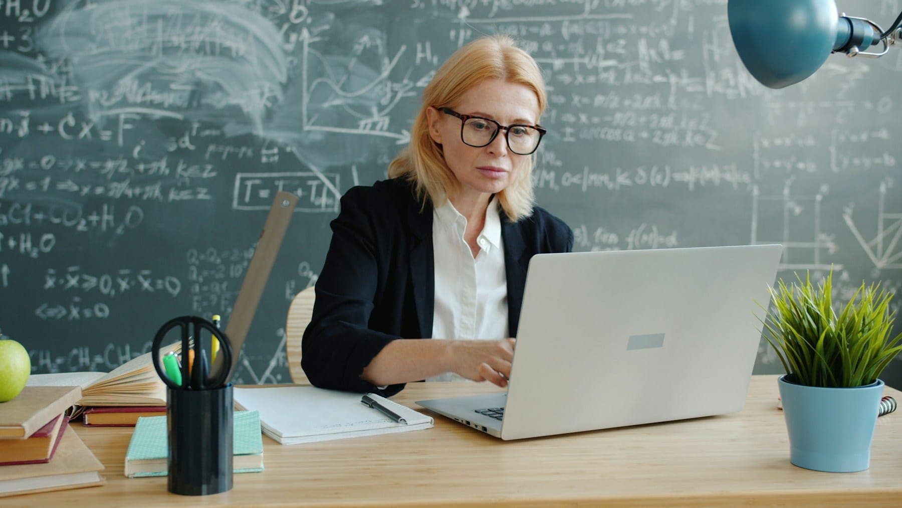 A teacher working on a laptop at a desk in front of a chalkboard covered in handwritten equations.