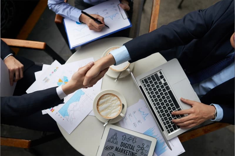 Two people in suits shaking hands over a café table covered with printed charts, coffee cups and a laptop, seen from above.