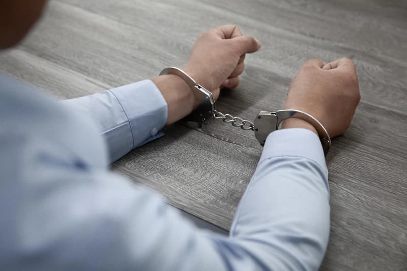 A man in a light blue shirt resting his handcuffed wrists on a wooden table.