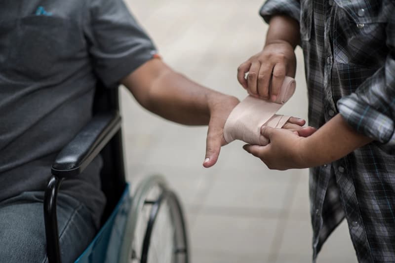 A person wrapping a bandage around the hand of someone seated in a wheelchair.