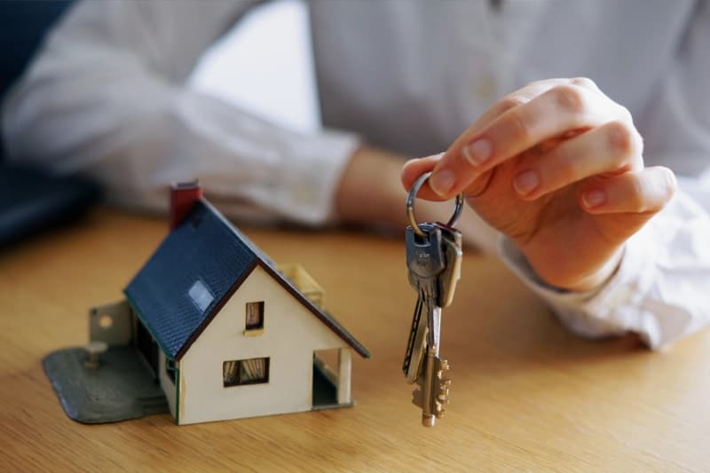 A hand holding a bunch of keys above a wooden table, beside a small model house.