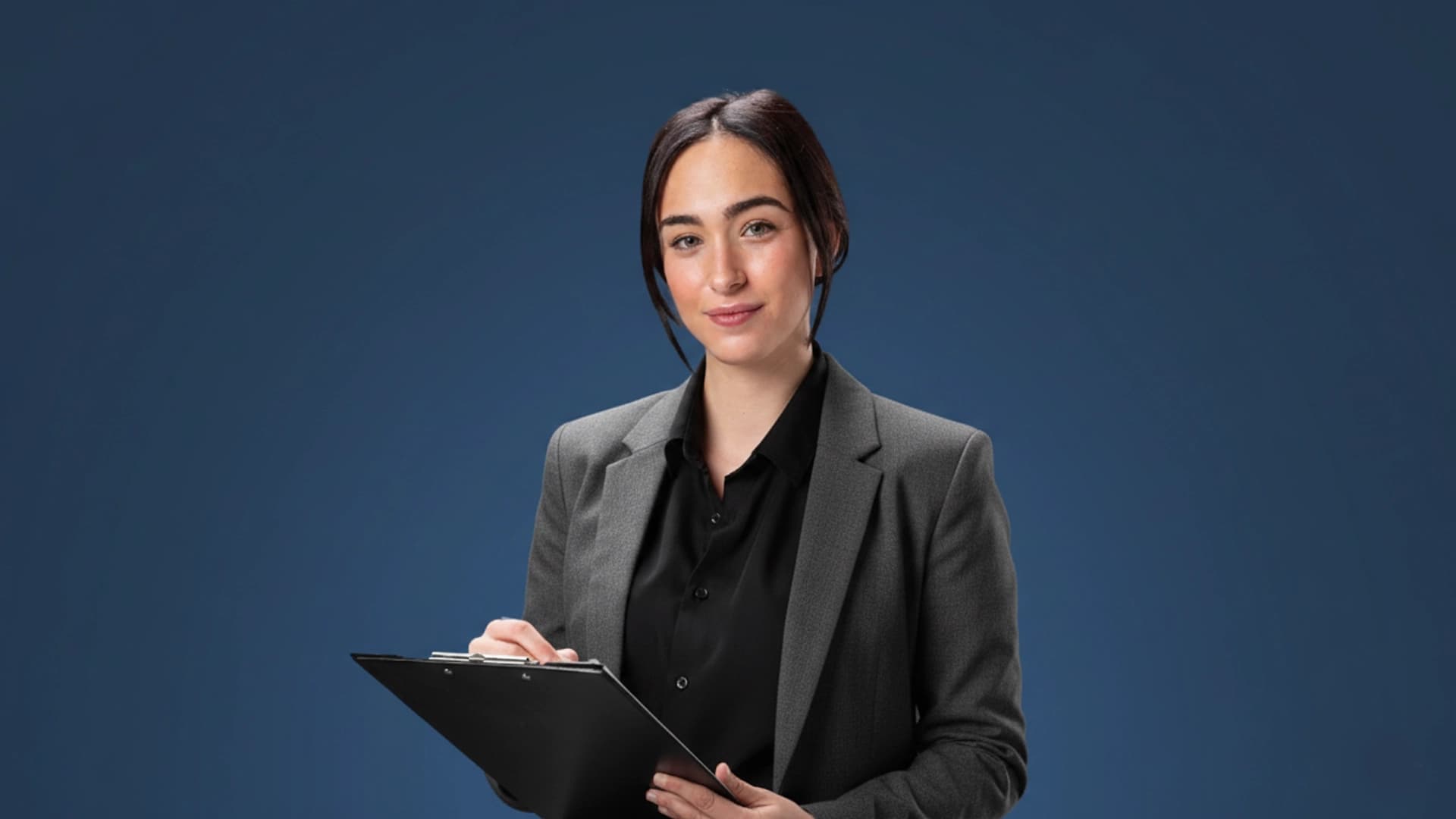 A woman in a grey blazer holding a clipboard and pen against a dark blue background.