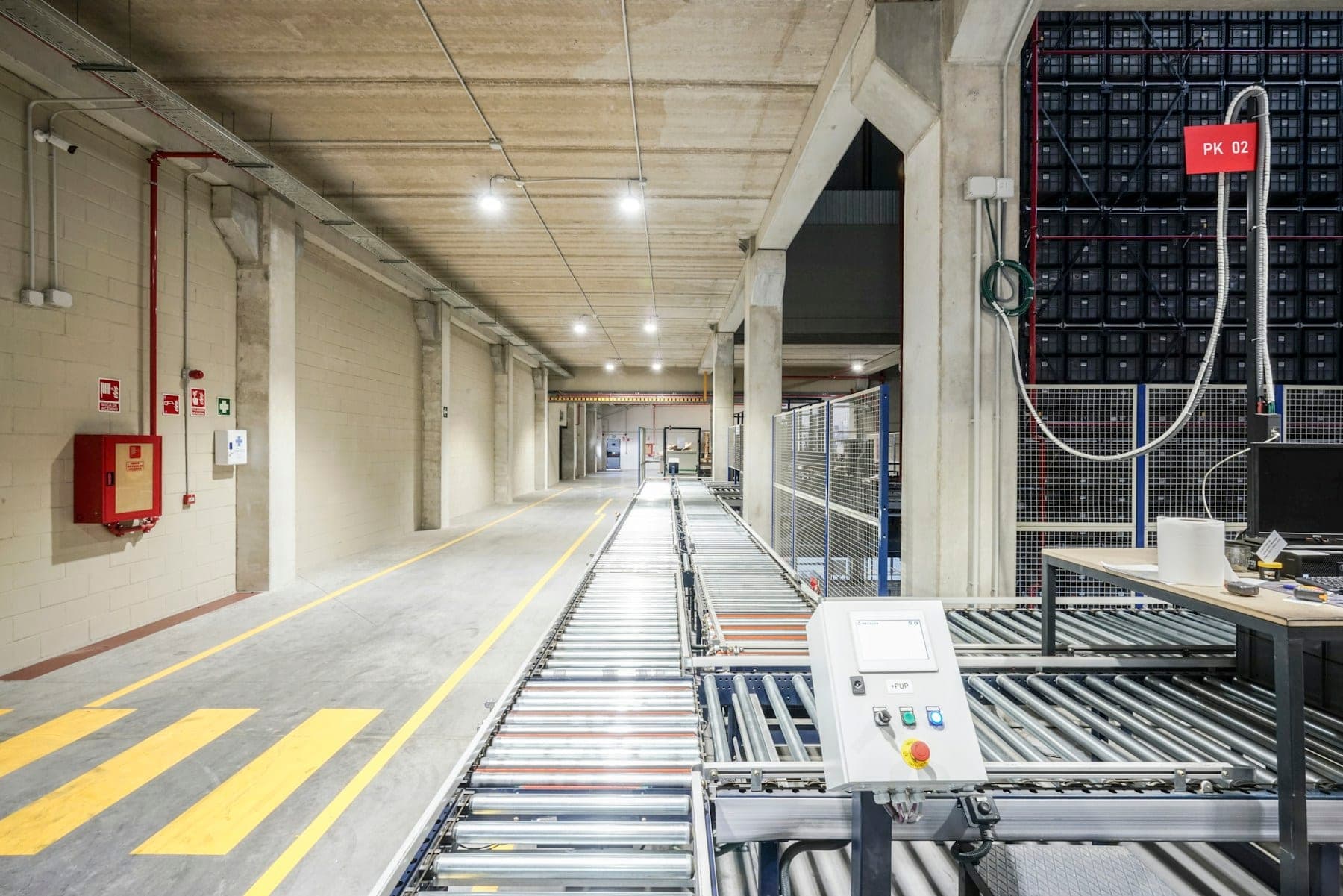 A conveyor line running the length of an automated distribution warehouse, past marked floor lanes and numbered picking bays.