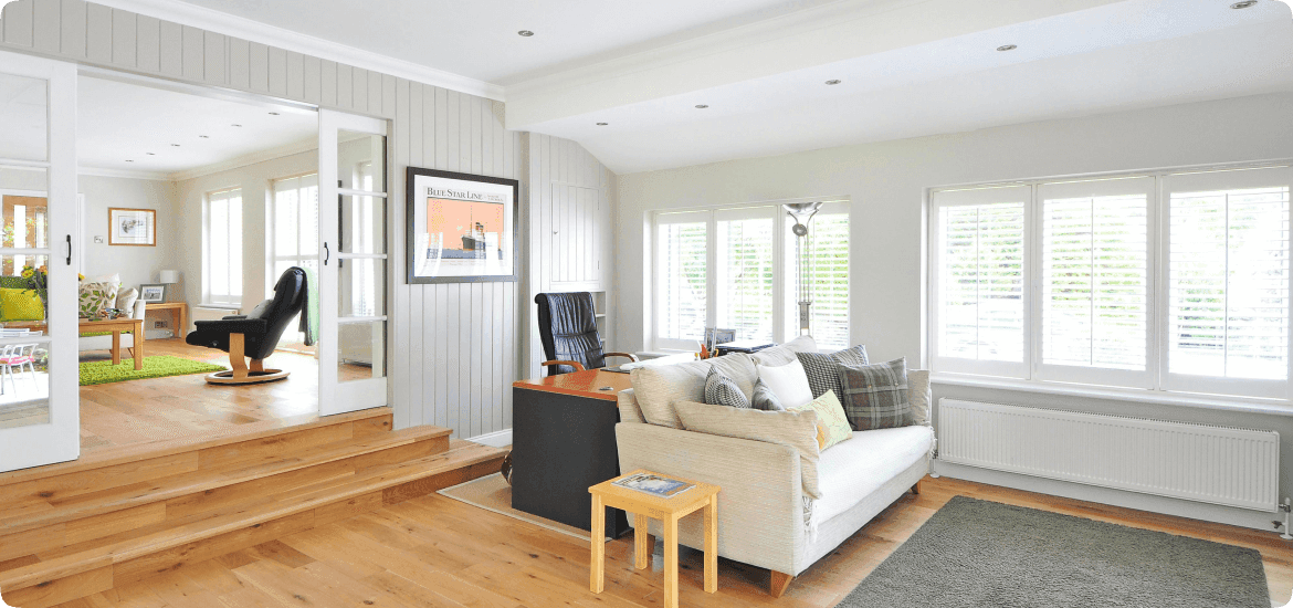 An open-plan living room with a wooden floor, a cream sofa and a desk, opening through glazed doors onto a raised second room.