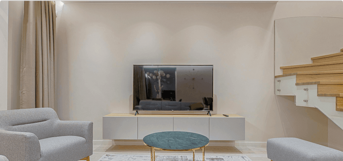A minimal living room with a wall-mounted television, grey armchairs and a round green marble table beside a floating wooden staircase.