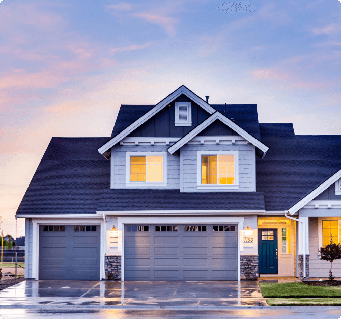 A blue-grey house at dusk with a teal front door and two garage doors, its windows glowing above a wet driveway.