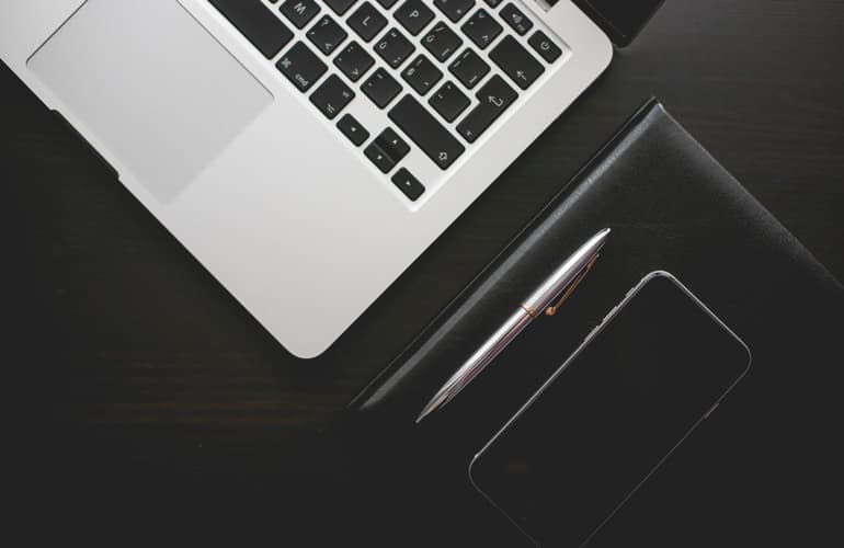 A laptop, a leather notebook, a pen and a phone arranged on a dark wooden desk, seen from above.