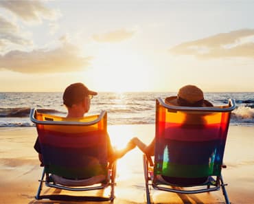 Two people holding hands in deck chairs on a beach, watching the sun set over the sea.