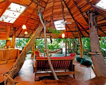 The open interior of a timber lodge with a high thatched roof, a cushioned bench between tree-trunk posts.
