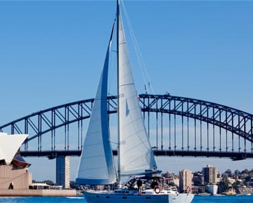 A sailboat under full sail crossing a harbour in front of a steel arch bridge, with a city skyline behind.