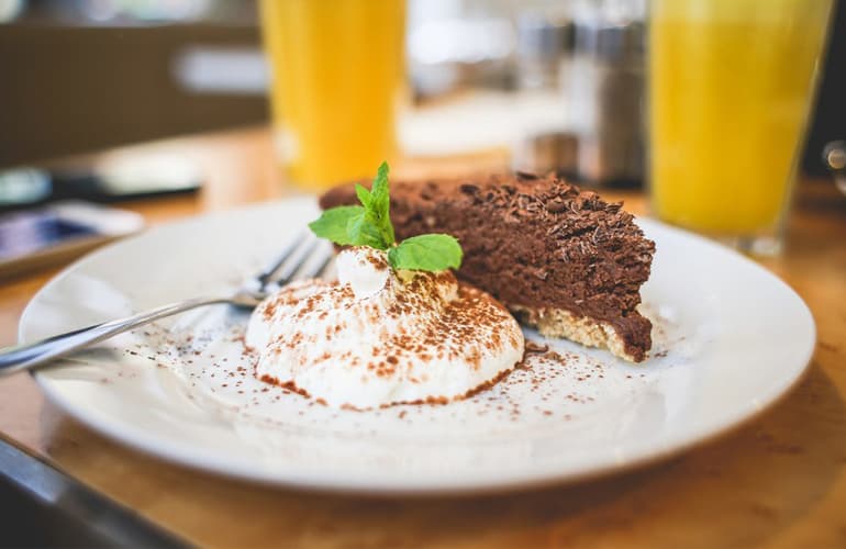 A slice of chocolate cake with cream and a mint sprig on a white plate, two glasses of juice behind it.