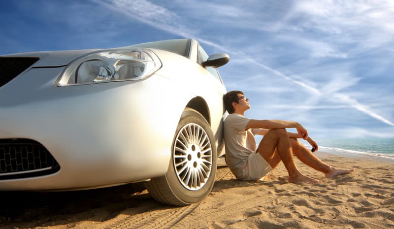 A man sitting on the sand leaning against the front wheel of a silver car on a beach, the sea behind him.
