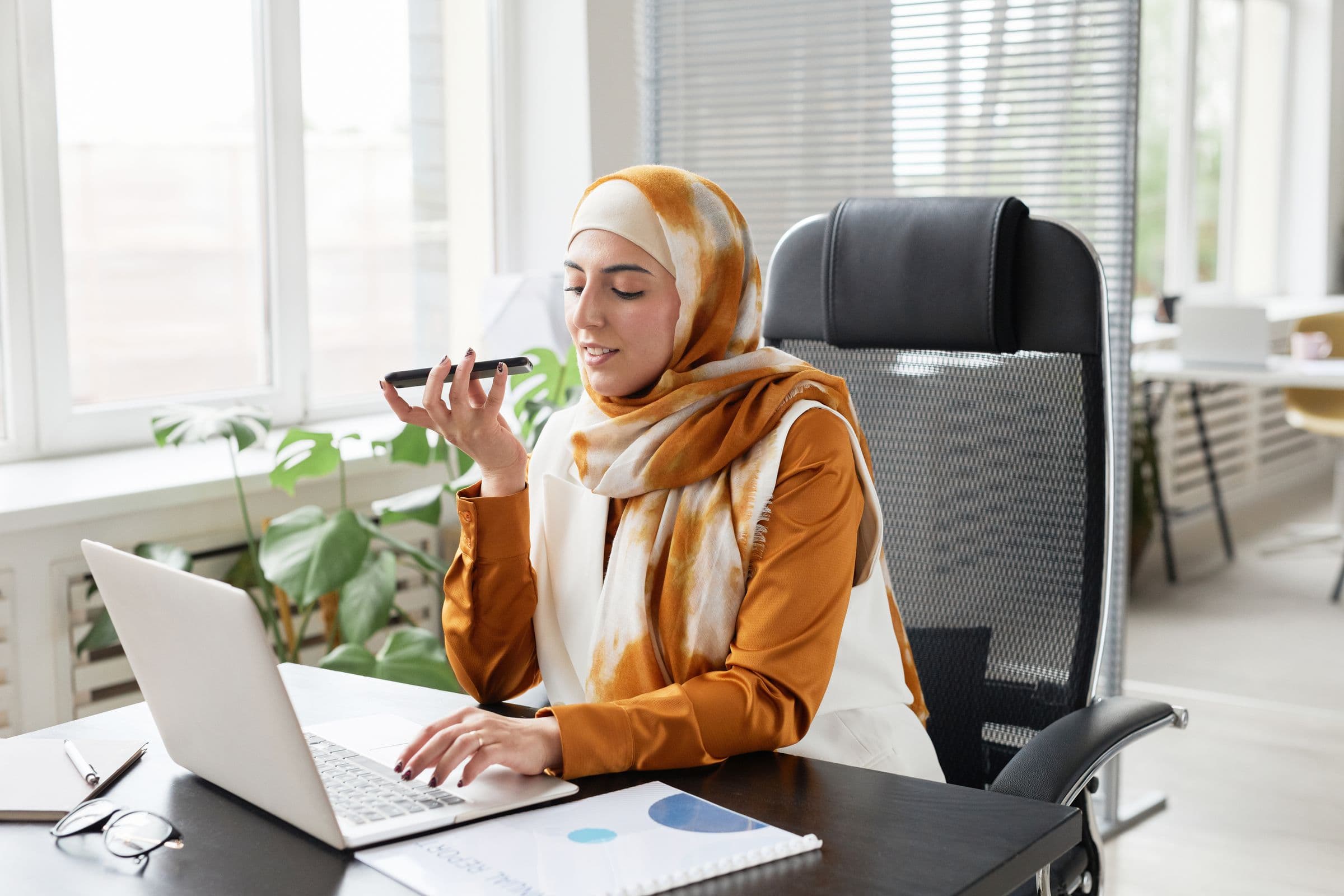Arab businesswoman using a phone and laptop to manage WhatsApp conversations for a Gulf business